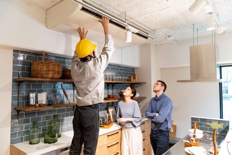 Inspector checking the functionality of an air conditioning unit in a kitchen while man and woman look on